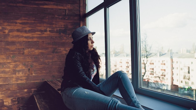 Jovem linda de chapéu e jaqueta de couro sentada perto das janelas Retrato de mulher bonita olhando pela janela durante o dia