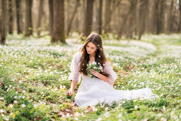 Jovem linda de vestido branco coleta prímulas Uma garota na floresta da primavera Um buquê de anêmonas brancas nas mãos