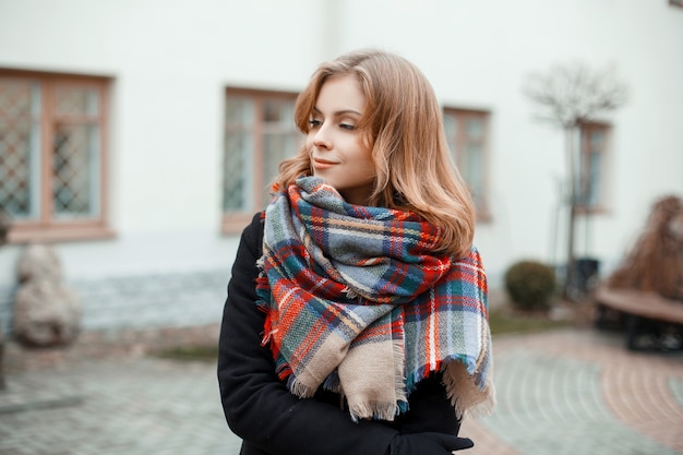 Jovem linda e doce mulher com um casaco quente de inverno da moda e um lenço de lã xadrez elegante fica em um dia quente de outono