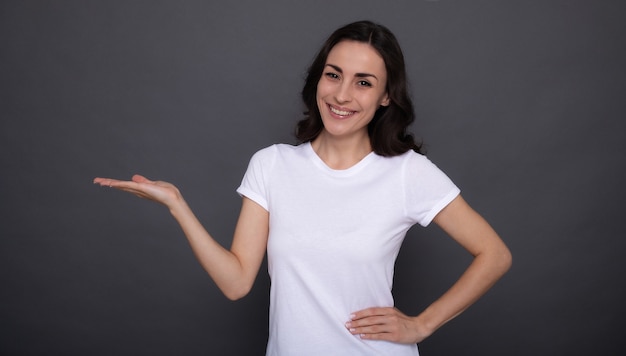 Jovem, linda e elegante mulher feliz em uma camiseta branca está posando e apontando para o fundo cinza