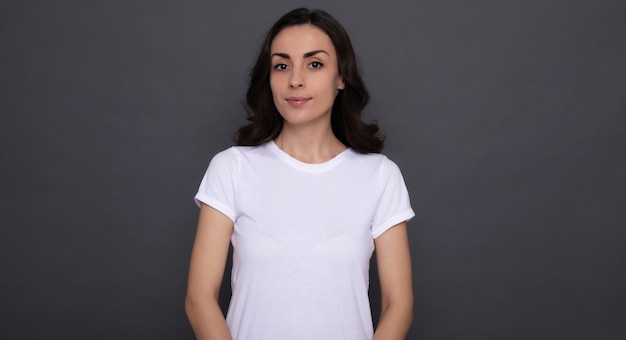 Jovem, linda e elegante mulher feliz em uma camiseta branca está posando no fundo cinza