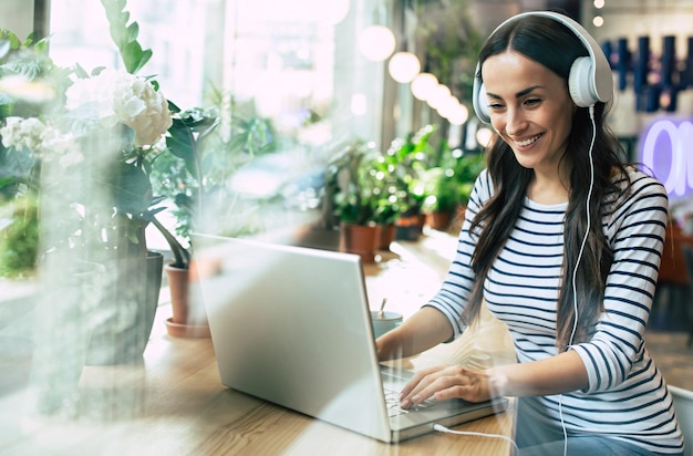 Jovem linda e feliz em fones de ouvido está ouvindo música no laptop no café ou no centro de trabalho