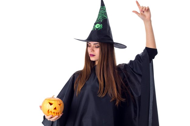 Jovem linda em traje preto de bruxa com chapéu preto segurando uma abóbora e olhando para ela