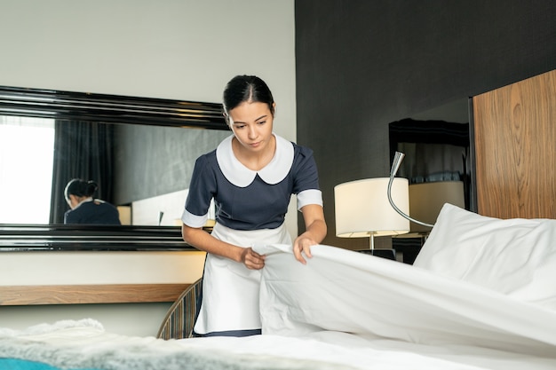 Foto jovem, linda empregada morena, colocando um cobertor branco limpo enquanto troca de cama em um dos quartos do hotel durante a preparação para novos hóspedes