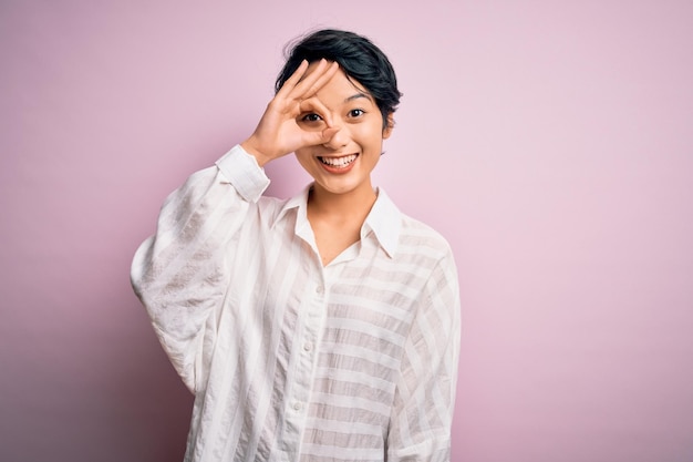 Jovem linda garota asiática vestindo camisa casual em pé sobre fundo rosa isolado fazendo okey gesto com mão sorrindo olho olhando por entre os dedos com cara feliz