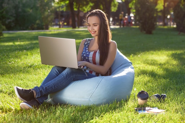 Jovem linda garota com laptop sentado em um saco de feijão no jardim ou parque na grama verde conceito de compras online