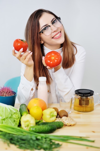 Jovem linda garota de óculos, segurando tomates vermelhos maduros na superfície de uma mesa com muitos legumes