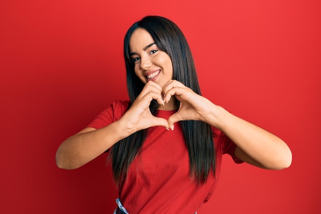 Jovem linda garota hispânica vestindo camiseta vermelha casual sorrindo apaixonada fazendo forma de símbolo de coração com as mãos. conceito romântico.