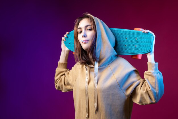 Jovem linda hipster elegante vestida com um capuz segura um skate nas mãos em um fundo de estúdio roxo come um pirulito