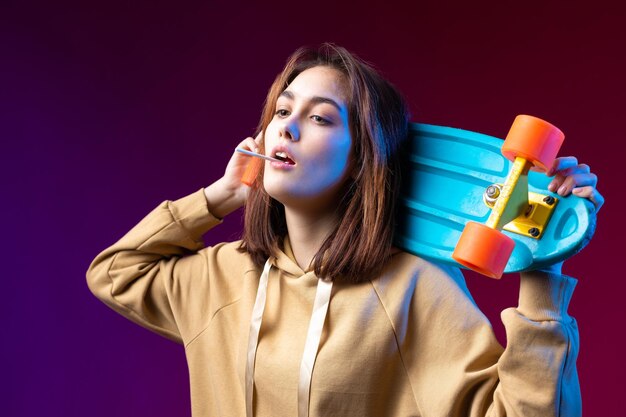 Foto jovem linda hipster elegante vestida com um capuz segura um skate nas mãos em um fundo de estúdio roxo come um pirulito