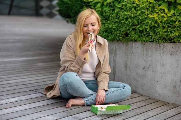 Jovem linda loira comendo sushi ao ar livre no terraço de madeira pelo edifício moderno na cidade Comida saborosa para viagem Garota tem intervalo para o almoço passando o tempo fora e comendo comida asiática Vida na cidade