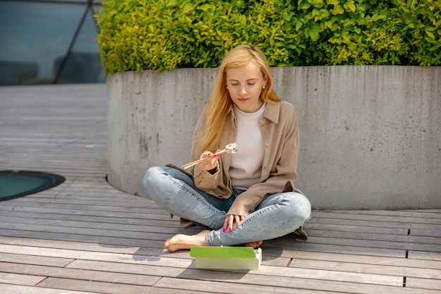 Jovem linda loira comendo sushi ao ar livre no terraço de madeira pelo edifício moderno na cidade Comida saborosa para viagem Garota tem intervalo para o almoço passando o tempo fora e comendo comida asiática Vida na cidade