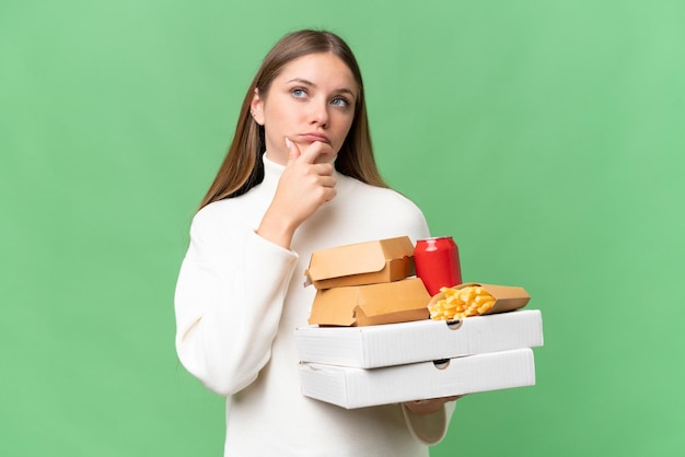 Jovem linda loira segurando comida para viagem sobre fundo isolado com dúvidas