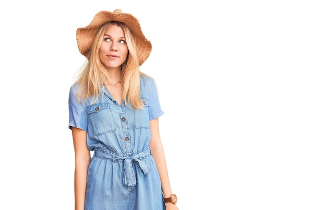 Jovem linda loira usando chapéu de verão e vestido sorrindo olhando para o lado e olhando para longe pensando