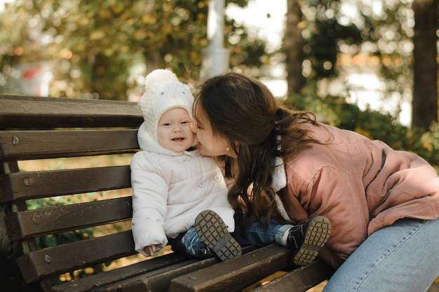 Jovem, linda mãe beija sua filha. feliz, mãe e bebê estão sentados em um banco no outono e sorrindo.