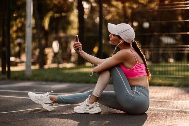 Foto jovem linda menina ouvindo música em fones de ouvido depois de uma prática esportiva no campo de esportes ao pôr do sol