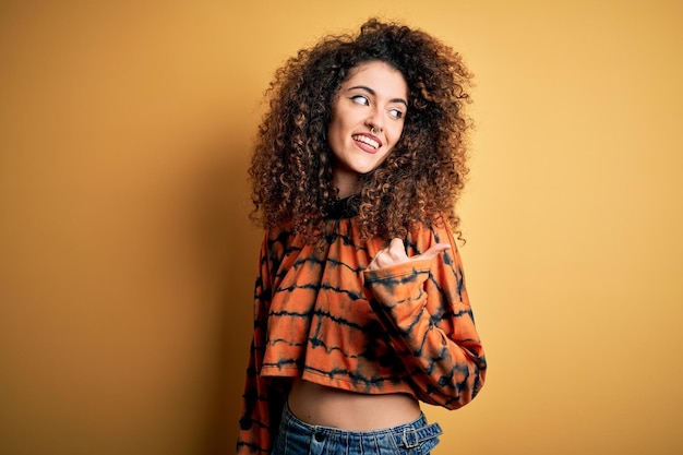 Jovem linda morena com cabelos cacheados e piercing vestindo camiseta casual sorrindo com cara de feliz olhando e apontando para o lado com o polegar para cima