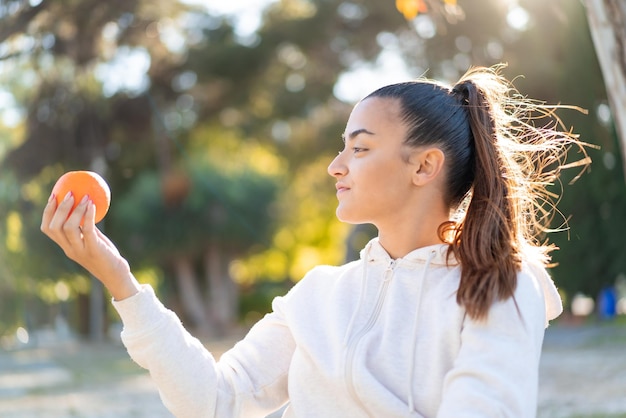 Jovem linda morena segurando uma laranja ao ar livre com expressão feliz