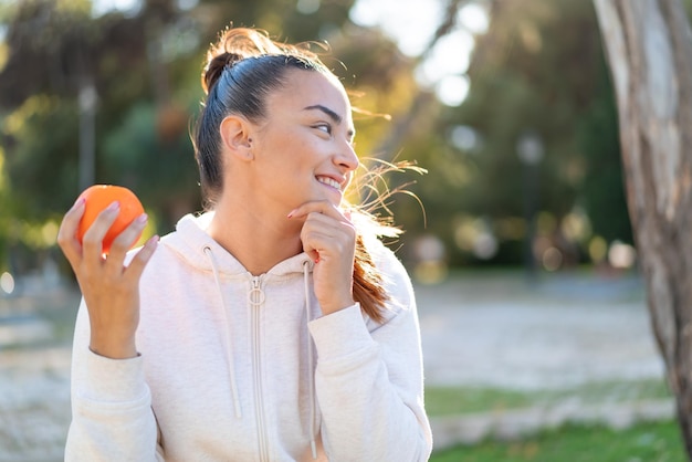 Jovem linda morena segurando uma laranja ao ar livre pensando em uma ideia e olhando de lado