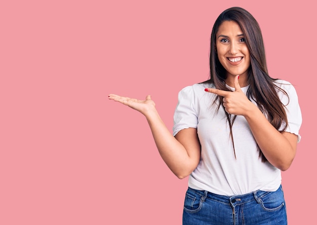 Jovem linda morena vestindo camiseta casual espantada e sorrindo para a câmera enquanto apresenta com a mão e aponta com o dedo