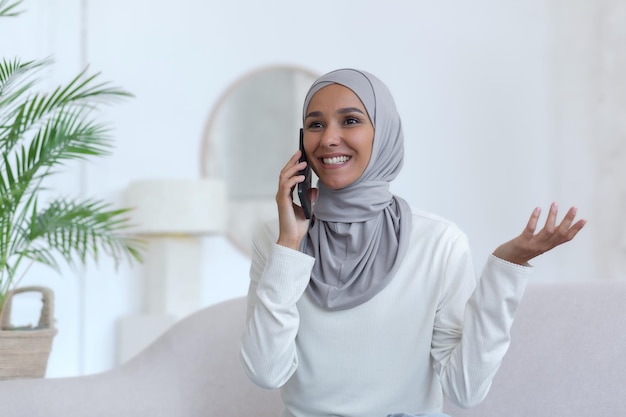 Jovem linda muçulmana em hijab em casa falando ao telefone mulher na sala sorrindo e