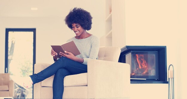 Jovem linda mulher afro-americana sentada em frente à lareira em casa em um dia frio de outono e lendo livro