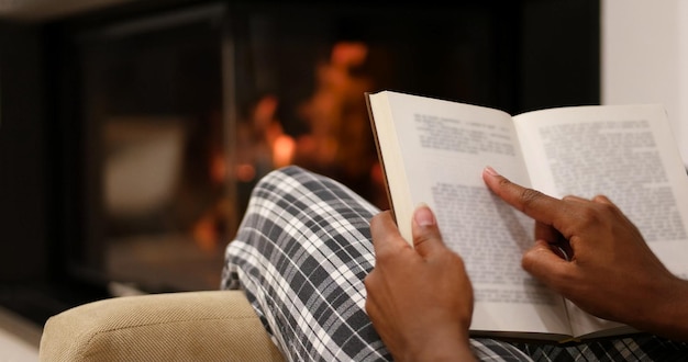 Jovem linda mulher afro-americana sentada em frente à lareira em casa em um dia frio de outono e lendo livro