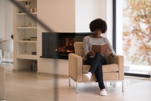 Jovem linda mulher afro-americana sentada em frente à lareira em casa em um dia frio de outono e lendo livro