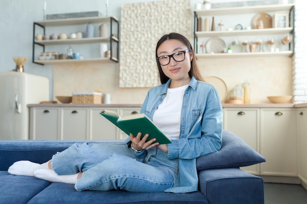 Jovem linda mulher asiática em roupas jeans e óculos sentados no sofá em casa ele segura um verde
