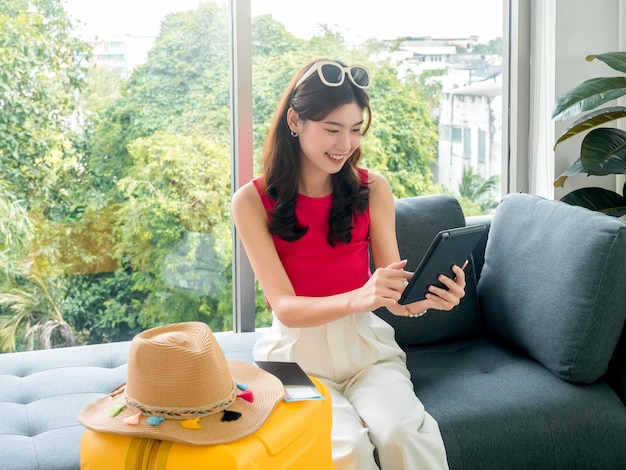 Jovem linda mulher asiática feliz com óculos de sol usando computador tablet digital com passagem aérea de passaporte e chapéu de praia na mala amarela sente-se no sofá pronto para viajar no conceito de férias de verão