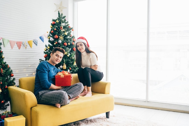 Jovem linda mulher asiática feliz usando um chapéu de Papai Noel surpreende o namorado com um presente em casa com uma árvore de Natal ao fundo Comemore o conceito do boxing day Foto com espaço de cópia