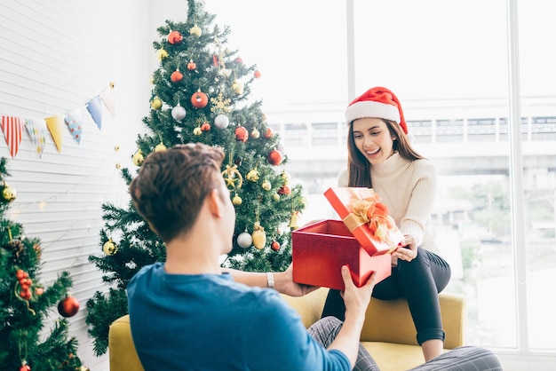 Jovem linda mulher asiática feliz usando um chapéu de Papai Noel surpreende o namorado com um presente em casa com uma árvore de Natal ao fundo Comemore o conceito do boxing day Foto com espaço de cópia