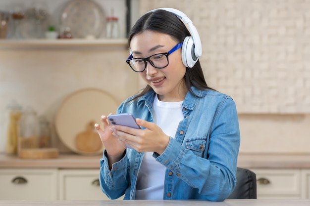 Jovem linda mulher asiática ouvindo música no telefone sentada em casa na cozinha vestindo