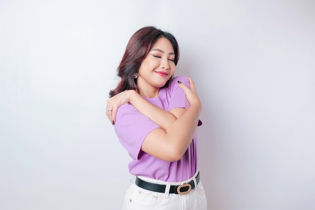 Jovem linda mulher asiática vestindo uma camiseta roxa lilás sobre fundo branco se abraçando feliz e positiva sorrindo confiante Amor próprio e autocuidado