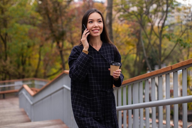Jovem linda mulher casual de casaco com café para ir alegremente falando no celular no parque