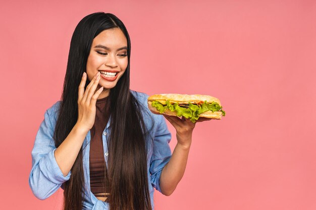 Jovem linda mulher chinesa asiática comendo sanduíche ou hambúrguer grande com satisfação Garota gosta de conceito de dieta para viagem de hambúrguer saboroso isolado sobre fundo rosa