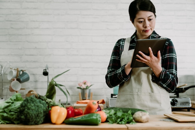 jovem linda mulher coreana asiática vestindo camisa e avental usando tablet digital na cozinha de madeira vintage para receita. esposa elegante assistindo touch pad em pé no local de cozimento pesquisando on-line.