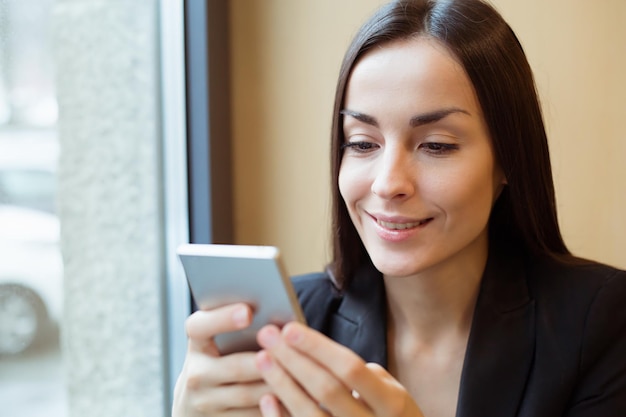 Jovem linda mulher de negócios sorridente em uma jaqueta preta trabalhando no telefone ou usando a Internet sentada em uma mesa em um café