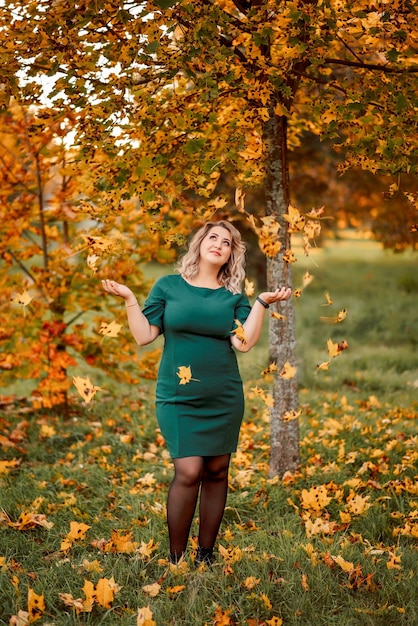 Jovem linda mulher de tamanho grande em um vestido verde joga folhas em um parque de outono