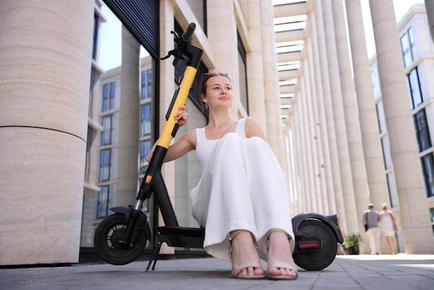 Jovem linda mulher feliz muito atraente adolescente está montando eletro scooter na cidade no dia ensolarado de verão em roupas brancas sorrindo sentado no transporte de compartilhamento de eco elétrico