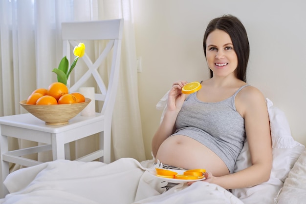 Jovem linda mulher grávida comendo laranjas sentado na cama em casa. Comida saudável, vitamina C natural