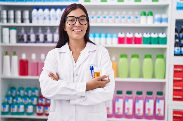 Jovem linda mulher hispânica farmacêutica de pé com os braços cruzados gesto segurando garrafa de pílulas na farmácia