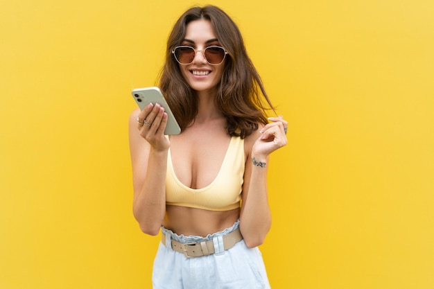 Jovem linda mulher sorridente com telefone celular Mulher despreocupada posando na rua perto da parede amarela Modelo positivo ao ar livre em óculos de sol Feliz e animado