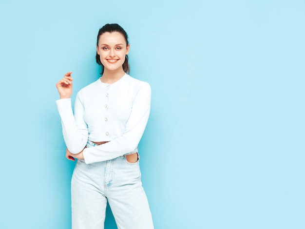 Jovem linda mulher sorridente em jeans da moda verão e roupas de cima Mulher despreocupada sexy posando perto da parede azul no estúdio Modelo morena positiva se divertindoAlegre e feliz