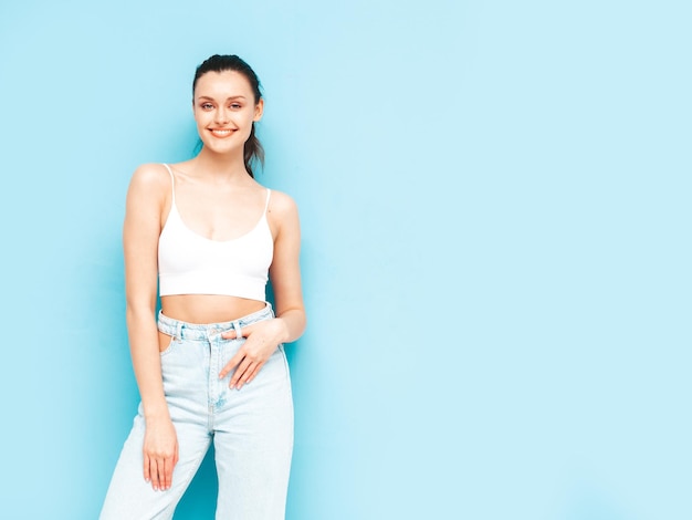 Foto jovem linda mulher sorridente em jeans da moda verão e roupas de cima mulher despreocupada sexy posando perto da parede azul no estúdio modelo morena positiva se divertindoalegre e feliz