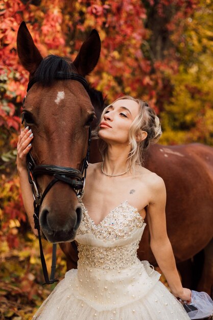 Jovem linda noiva em um exuberante vestido branco feliz caminhando com um cavalo na floresta de outono