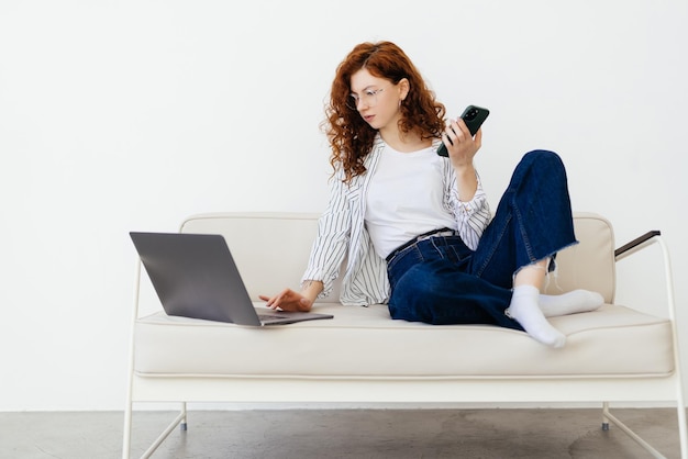 Jovem linda ruiva sentada no sofá na sala de estar e fazendo compras online no celular e laptop