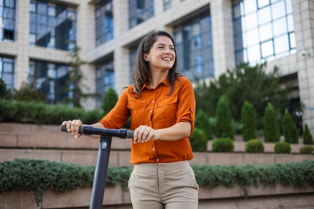 Jovem linda sorri e anda de scooter elétrica para trabalhar ao longo de edifícios de escritórios Conceito de transporte ecológico