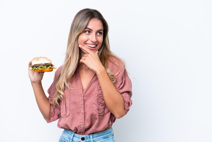 Foto jovem linda uruguaia segurando um hambúrguer isolado no fundo branco, pensando em uma ideia e olhando de lado