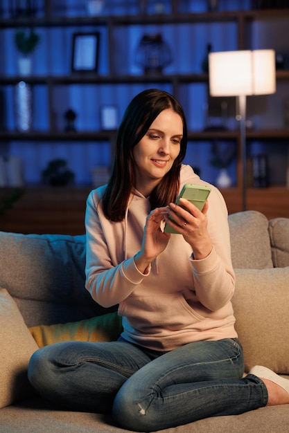 Jovem linda usando aplicativos de celular navegando nas mídias sociais da internet olhando para smartphone e sorrindo lazer em casa à noite iluminação cinematográfica